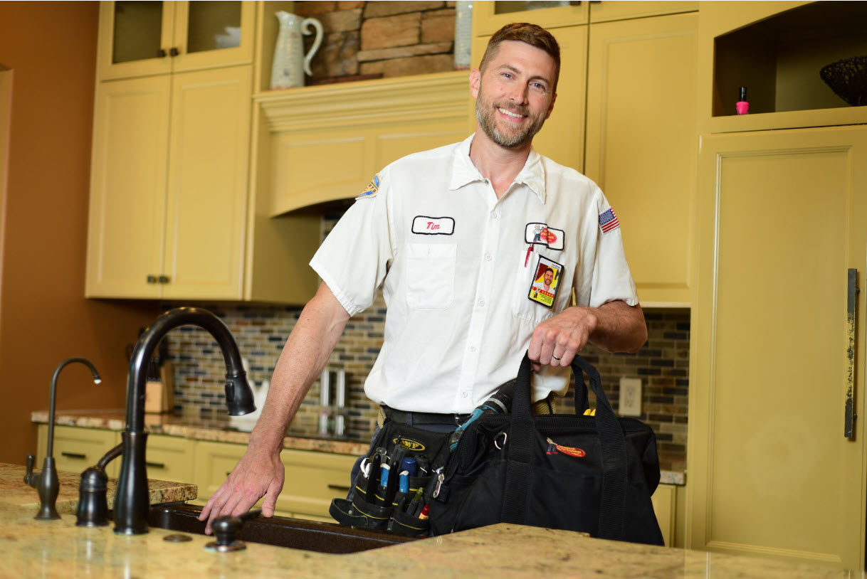 Service Professor technician smiling and holding tool bag while standing next to a kitchen sink.