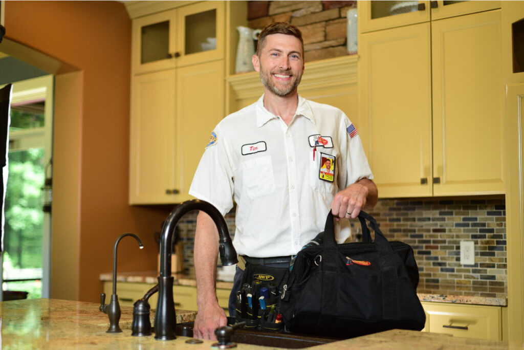 Smiling Service Professor Technician standing in front of kitchen sink, holding black tool bag.