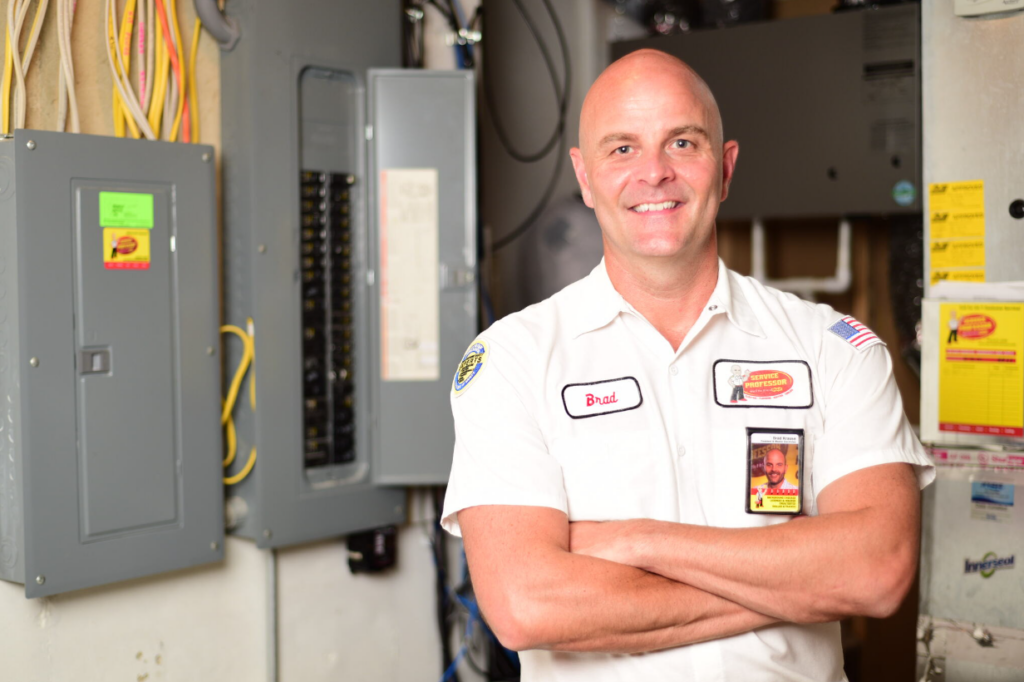 Service Professor electrician smiling with arms crossed in front of him, in front of an electrical panel.